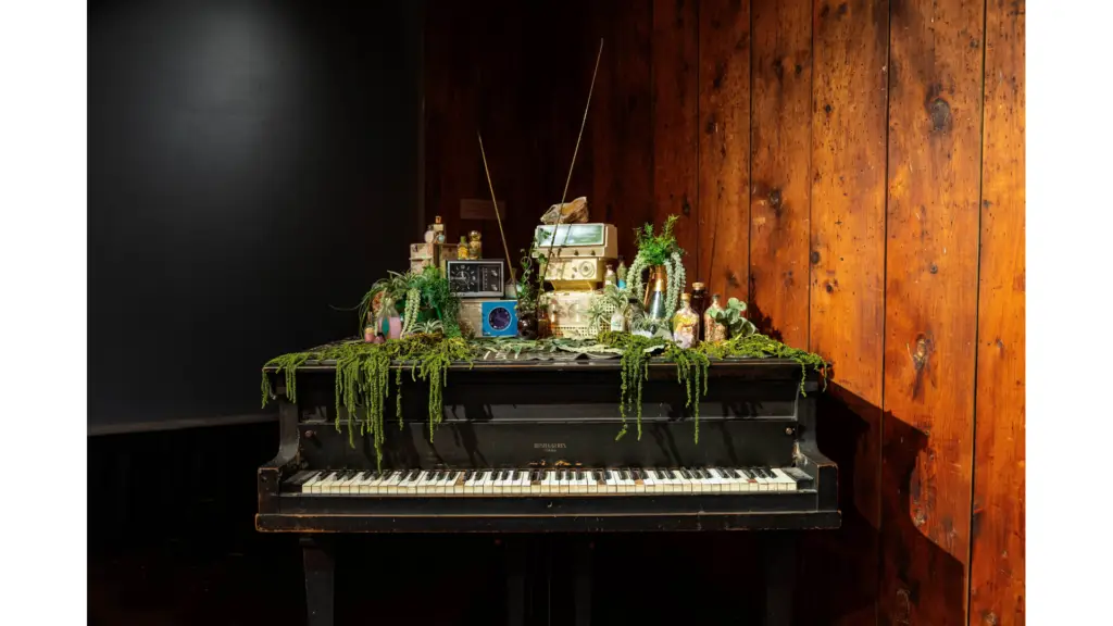 An installation of clocks, radios, and plants, on top of a piano.