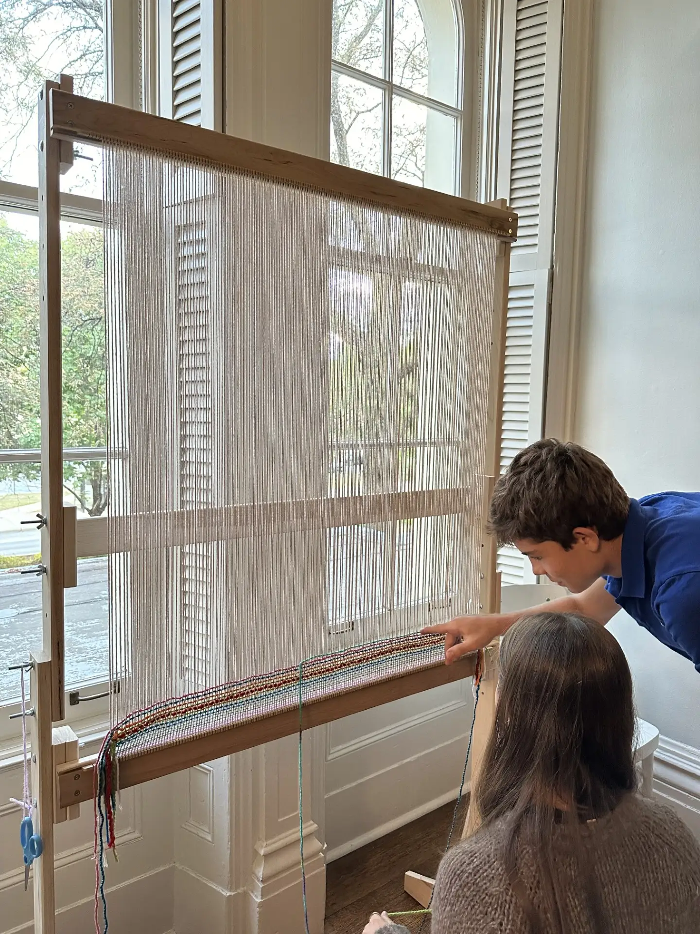 Two young visitors look at a large standing loom in front of a window of Hull-House.