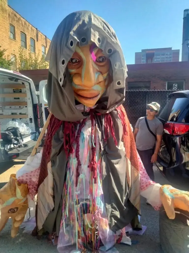 A large-scale puppet wears a cloak and hood, its face and large hands visible.