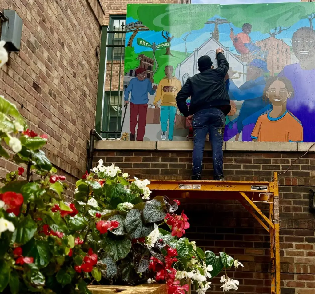 A figure wearing a cap stands on a scaffold with back to the camera, working on a mural on a brick wall.