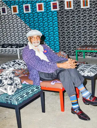 Robert Paige, wearing a purple jacket, hat, and striped socks, sits in a chair in front of patterned wallpaper.