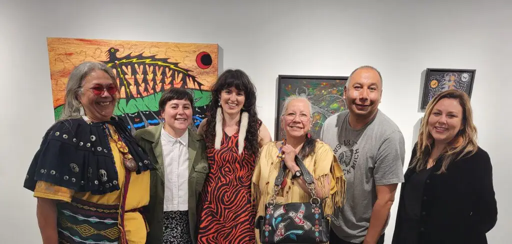 Four artists and two curators pose together in front of an gallery wall.