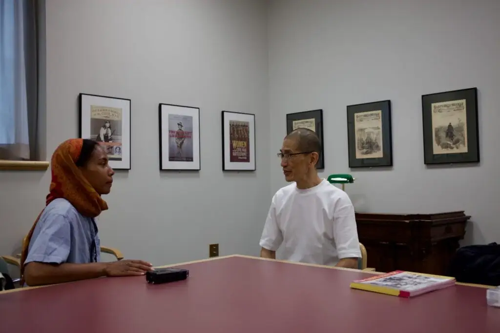 A photograph of Mary Lawson interviewing Akito Tsuda. They sit at a big red table with photographs hung on a wall in the background.