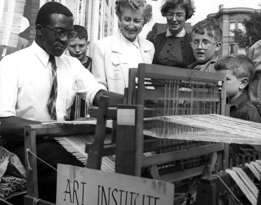 A vintage photo of an artist seated at a weaving loom. A group of adults and children look on.