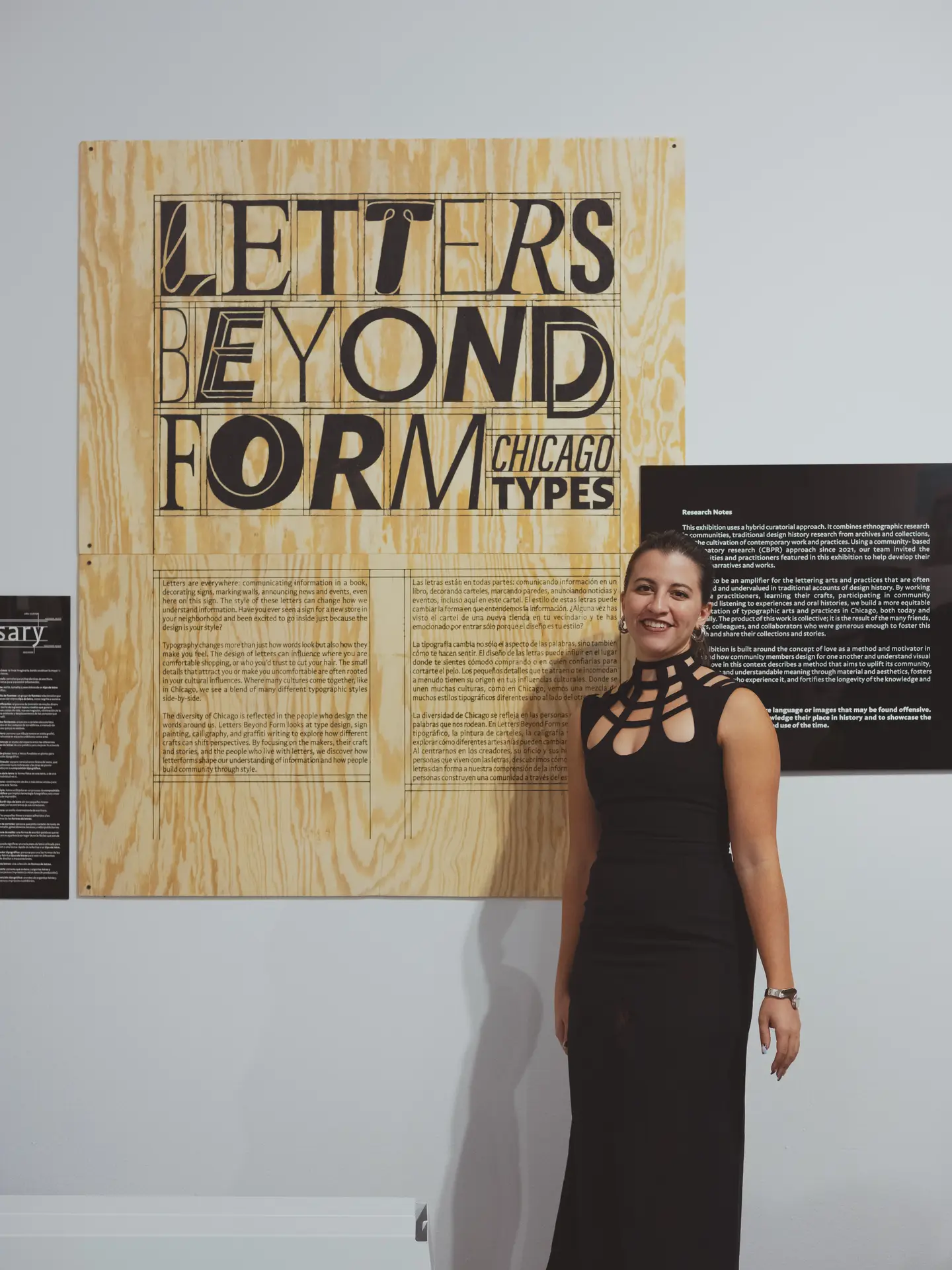 Curator Amira Hegazy, wearing a long black dress and hair pulled back, stands in front of the opening exhibition text panel for "Letters Beyond Form."