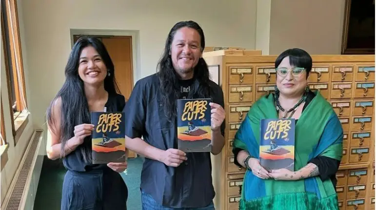 Jim Terry, with long hair and wearing a t-shirt, stands with Haku Blaisdell on his right and Analú Maria Lopez on his left. They each hold a copy of "Paper Cuts."