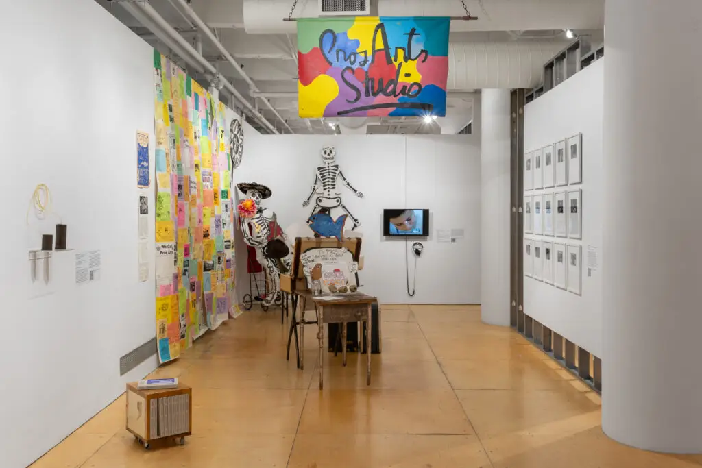 Exhibition installation view showing handmade objects including a large skeleton puppet. A sign above reads "Pros Arts Studio."