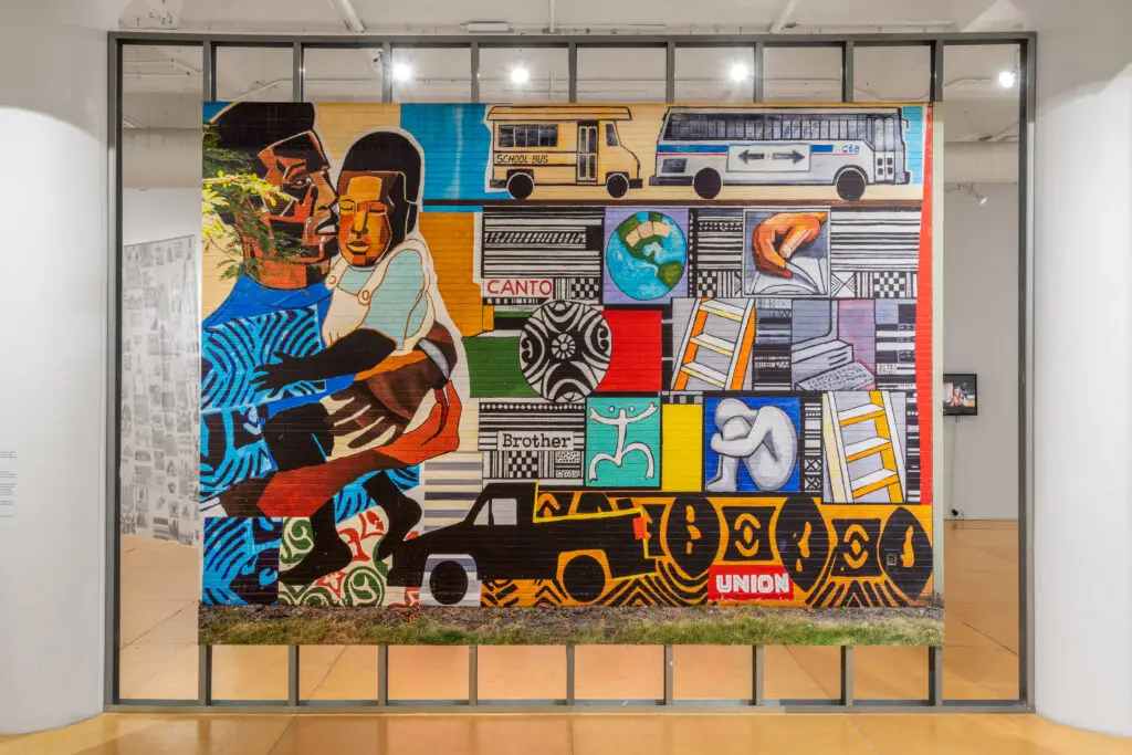 Gallery installation view of a colorful mural depicting a Black man holding a child, a car, two busses, and a variety of other objects.