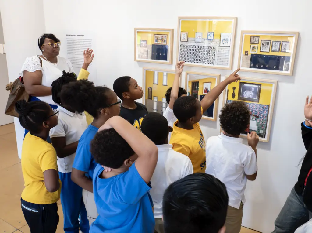 A group of young students in a gallery space looking at the exhibition and pointing to objects on a gallery wall.