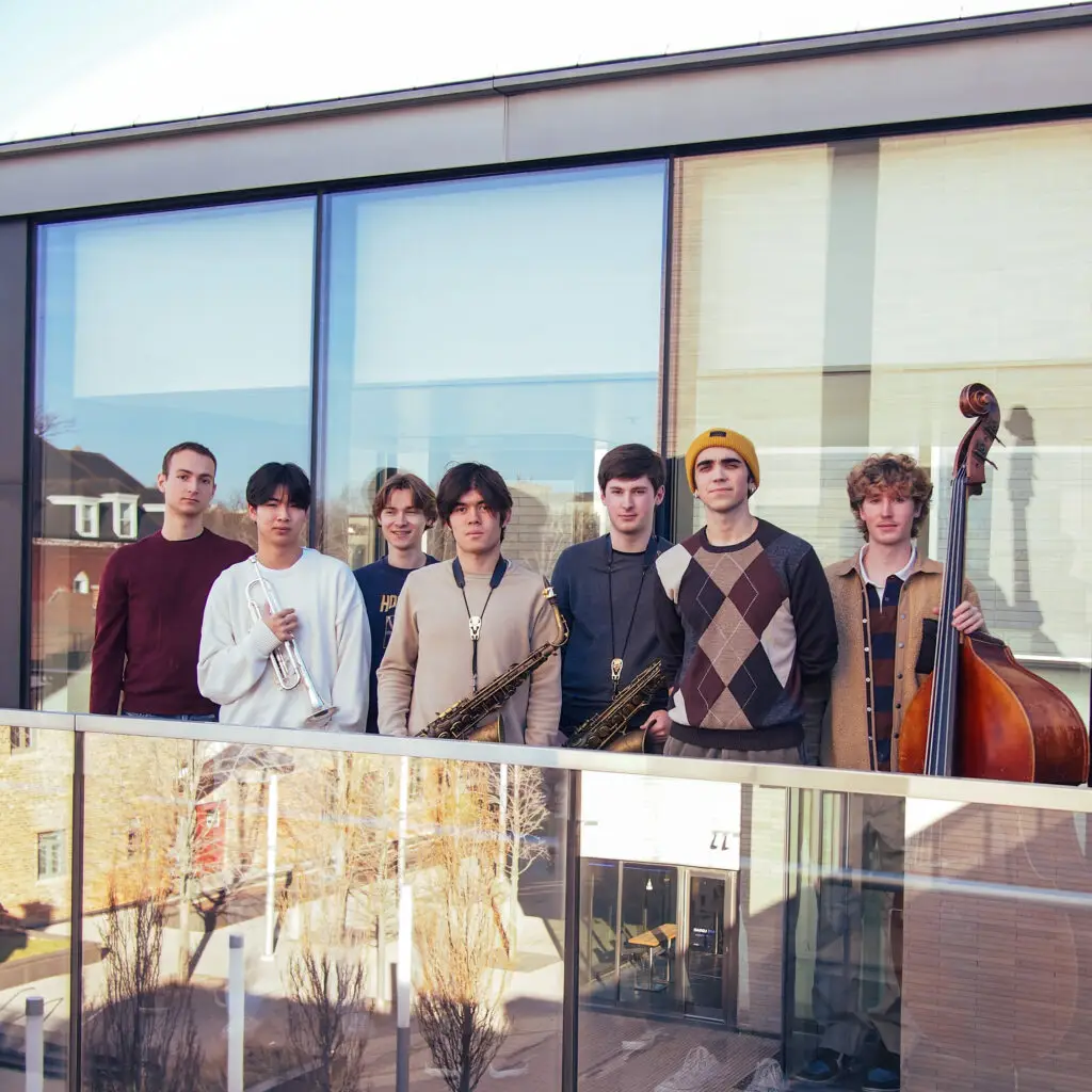 A group of young musicians stands on a balcony, several holding instruments.