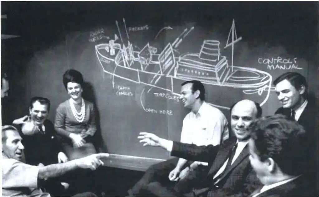 A vintage photo of a group of coworkers sitting in front of a blackboard with a picture of a ship on it.