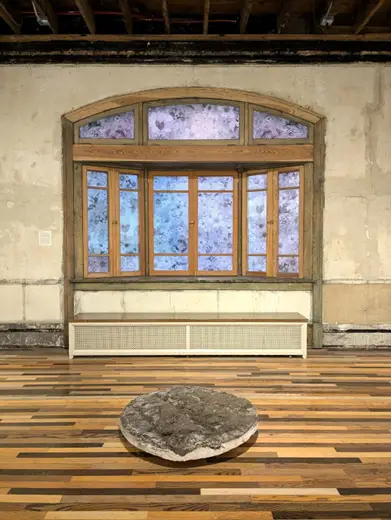 A large, flat, round sculpture sits on a wooden floor in front of a window covered with a floral patterned film.